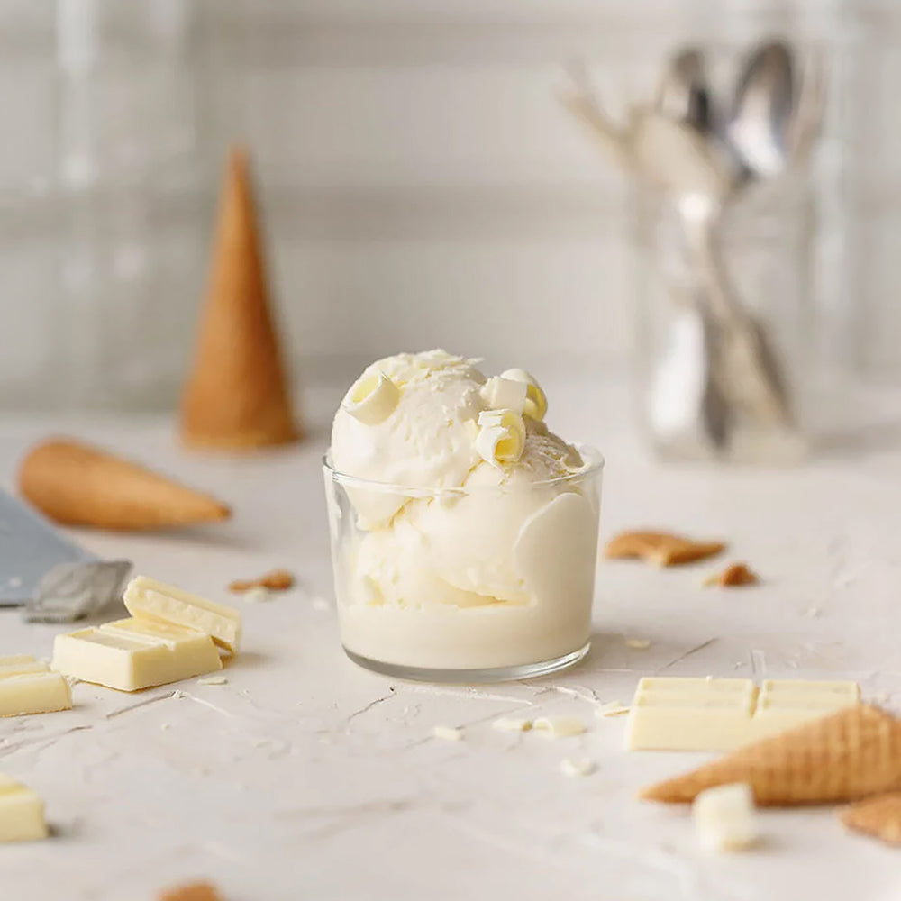 EVS07064 White Chocolate Flavour Paste for Gelato & DessertsPasta CioccobiancoWhiteChocolateFlavourPasteforGelato Glass bowl of vanilla ice cream with white chocolate pieces on a light surface.
