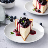 Slice of cheesecake with blueberry sauce on a white plate, surrounded by blueberries.