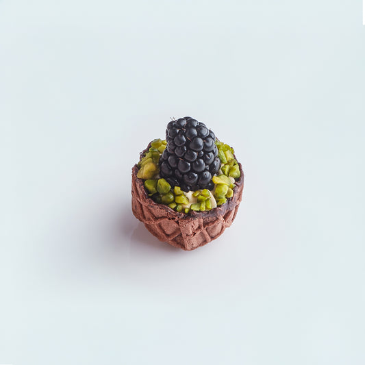 Small dessert with a chocolate base, green pistachios, and a black berry on a light blue background